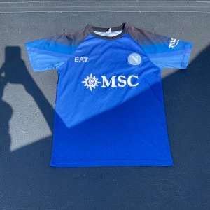 Napoli Maradona #10 fotbollströja EA7 - Snygg blå Napoli fotbollströja från EA7 med Maradona och nummer 10 på ryggen. Tröjan har korta ärmar, svarta och blå detaljer på axlarna och ett diskret mönster med Napoli-loggor över hela tyget. MSC-logga på bröstet. Perfekt för dig som älskar fotboll och klassiska spelare.