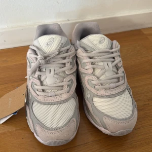 Gråvita sneakers från Asics (nypris 1800) - Snygga Asics sneakers i ljusgrått och vitt med meshpaneler och detaljer i mocka. Skorna har en chunky sula, klassisk snörning och ikoniska Asics-loggan på sidan. Perfekta för dig som gillar sportig och trendig stil.
