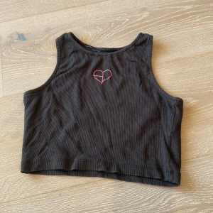 Svart croppad linnetopp BLACKPINK - Säljer en svart croppad linnetopp från BLACKPINK med ribbad struktur. Toppen har ett broderat rosa hjärta framtill och är ärmlös med hög halsringning. Perfekt för dig som gillar K-pop och vill ha en cool och bekväm topp till sommaren.