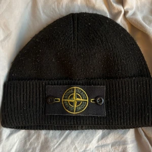 Svart stickad mössa Stone Island - Svart ribbstickad mössa från Stone Island med klassisk logotyp-patch framtill, fäst med två svarta knappar. Snygg och enkel design som passar till streetwear och håller dig varm under kalla dagar.