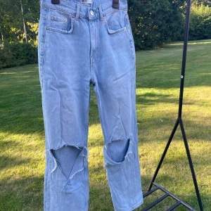 Ljusblå ripped jeans från Gina Tricot - Säljer ett par ljusblå jeans från Gina Tricot med stora slitningar över knäna. Klassisk femficksmodell i rak passform och hög midja. Jeansen är i denim och har en avslappnad vibe, perfekta för en cool streetstil. De är lite nedtrappade längst ned