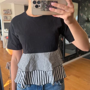 Svart topp med volang och ränder - Säljer en svart topp med kort ärm och volang nertill i svartvitt randigt tyg. Toppen har en cool kontrast mellan den enfärgade överdelen och den mönstrade volangen. Perfekt till jeans för en chill men snygg look.