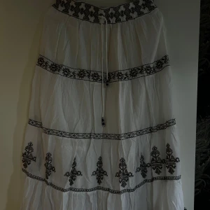 Vit långkjol med broderade detaljer! - Säljer en vit långkjol med svarta broderade mönster och snörning i midjan. Kjolen har broderier och är gjord i ett lätt, luftigt material som passar perfekt för varma dagar. 