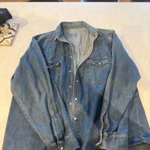 Jeansskjorta från Tezas i XL - Snygg jeansskjorta från Tezas i klassisk blå denim. Skjortan har två bröstfickor med lock och tryckknappar, markerad söm på ryggen och lång ärm. Väldigt avslappnad och pösig i modellen. Köpt på second hand för 700 kr, aldrig kommit till användning. Begär 250 kr