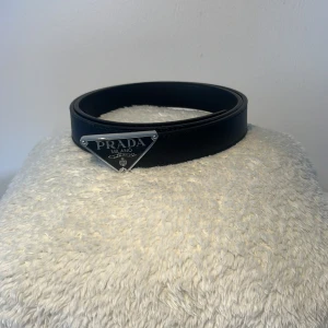 Svart skärp från Prada med silverdetalj - Snyggt svart bälte från Prada i skinn med en ikonisk trekantig silverfärgad metallplatta framtill där det står 'PRADA MILANO'. Bältet är stilrent och har klassisk design som passar till många olika outfits. Perfekt accessoar för att lyfta din look.