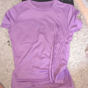 Lila t-shirt med snörning - Säljer en lila t-shirt med korta ärmar och cool snörningsdetalj på sidan. T-shirten har en rund halsringning och är gjord i mjukt material som känns skönt mot huden. Perfekt för dig som gillar trendiga och färgglada plagg. (Lila är mörkare än på bild)