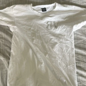 Vit t-shirt med strasstext från Bvaldi - Vit t-shirt från Bvaldi i bomull med korta ärmar. På bröstet finns VB i strass och på ryggen står det 'NEED MONEY FOR PORSCHE' i glittriga stenar. Enkel och clean passform, perfekt för dig som gillar statement-plagg.