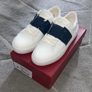 Valentino Garavani open mörk blå - Snygga vita sneakers från Valentino Garavani med marinblå detalj över snörningen och guldfärgad logga på plösen. Skorna är tillverkade i skinn och har en platt sula med mönstrad undersida. Perfekt för dig som gillar stilrena och lyxiga sneakers med unik design.