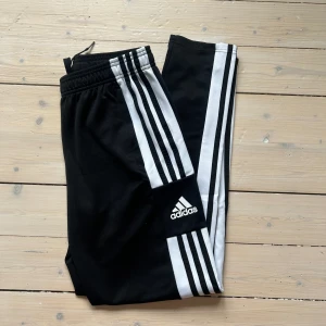 Adidasbyxor - Svarta (äkta) Adidas byxor med klassiska vita ränder längs sidorna och Adidas-logga på benet. Byxorna har resår i midjan och är gjorda i ett mjukt syntetmaterial (Polyester). I mycket fint skick. Skulle säga att det passar en XS-S