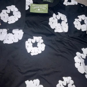 Svart t-shirt med vita blommor Denim Tears - Svart t-shirt från Denim Tears med stora vita blommönster över hela plagget. Klassisk rund hals och korta ärmar. Materialet är mjuk bomull som känns skönt mot huden. Perfekt statement-plagg för dig som gillar coola prints och streetwear.