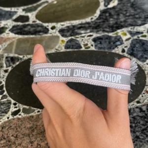 Grå Christian Dior J'Adior armband - Snyggt grått armband från Christian Dior med texten 'J'Adior' broderad i vitt. Armbandet har en mjuk, vävd struktur och avslutas med två stilrena tofsar. Perfekt accessoar för att lyfta din outfit med lite lyxig vibe.