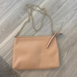 Rosa axelväska med kedjerem - Snygg rosa axelväska i skinnimitation med guldfärgad kedjerem och dragkedja upptill. Enkel och stilren design som passar till många outfits. Perfekt storlek för det viktigaste och lätt att bära över axeln.