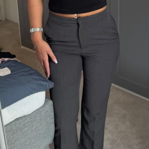 Mörkgrå kostymbyxor med bootcut - Snygga mörkgrå kostymbyxor med bootcut och hög midja. Byxorna har klassiska fickor bak och stängs med dragkedja och knapp framtill. Perfekta för en clean och stilren look. Materialet känns mjukt och följsamt.