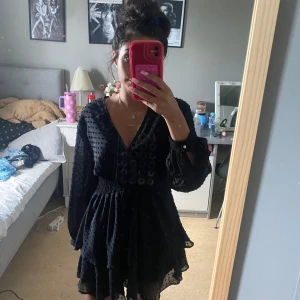 Svart omlottklänning med struktur från Zara - Svart omlottklänning från Zara med markerad midja och långärmade ärmar. Klänningen har en v-ringad hals och ett diskret prickigt strukturmönster i tyget. Perfekt för dig som gillar stilrena och trendiga plagg.