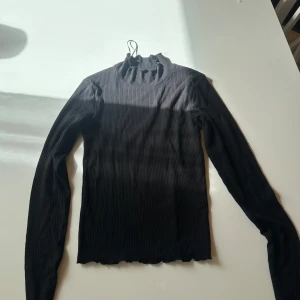 Svart ribbad polotröja från Bik Bok - Svart polotröja från Bik Bok i storlek XS. Tröjan är ribbad med lång ärm och har en hög krage med volangkant. Perfekt att styla med jeans eller kjol för en clean look. Materialet är mjukt och stretchigt, vilket gör den skön att bära hela dagen.