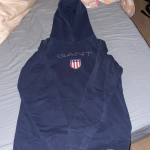 Marinblå hoodie från GANT - Säljer en marinblå hoodie från GANT med broderad logga och ett snyggt emblem på bröstet. Tröjan har huva, långa ärmar och är gjord i mjuk bomull. Perfekt för chill dagar och när du vill ha en klassisk look med streetvibe.