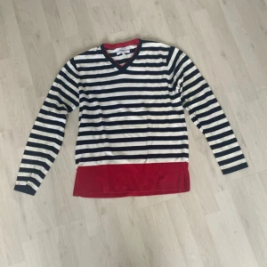 Randig tröja från GANT med röd kant - Snygg långärmad tröja från GANT i stretchig bomull. Tröjan har marinblå och vita ränder, röd kant nertill och vid v-ringningen. Perfekt för dig som gillar klassisk stil med en twist. Passar till jeans eller kjol för en chill look.