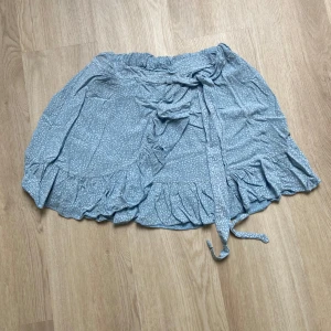 Blå omlottkjol med volang - Söt ljusblå kortkjol med små vita prickar, omlottmodell och knytband i midjan. Kjolen har volangkant längs nederdelen som ger en extra feminin touch. Perfekt för dig som gillar en avslappnad och trendig stil. Kan strykas innan någon köper så den inte blir så skrynklig. 