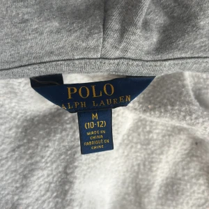 Grå barn hoodie från Polo Ralph Lauren - Klassisk grå hoodie från Polo Ralph Lauren med dragkedja framtill och huva. Tröjan har långa ärmar, känguruficka och diskret broderad logga på bröstet. Perfekt för chill dagar och enkel att matcha med jeans eller joggers.