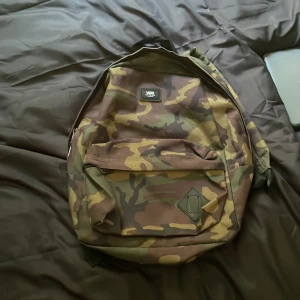Camouflage ryggsäck från Vans - Säljer en snygg ryggsäck från Vans med klassiskt camouflage-mönster i grönt, brunt och svart. Väskan har ett stort huvudfack, en framficka med dragkedja och justerbara axelremmar. Perfekt för skolan eller stan! Bara använd fåtal gånger! :3