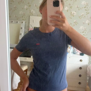 Blå Levi's t-shirt med logga - Snygg blå t-shirt från Levi's med klassisk röd logga på bröstet. Modellen är enkel med rund hals och korta ärmar, perfekt för en chill och stilren look. Materialet är mjuk bomull som känns skönt mot huden. Passformen är normal och sitter snyggt.