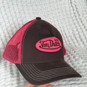 Brun och rosa Von Dutch keps - Skitsnygg oanvänd truckerkeps från Von Dutch i brunt och rosa med klassisk logga framtill. Kepsen har mesh-bakstycke i rosa för extra ventilation och vit kontrastsöm på skärmen. Perfekt accessoar för en cool streetstil.