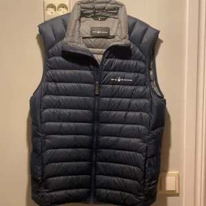 Mörkblå dunväst från Sail Racing - Säljer en snygg mörkblå dunväst från Sail Racing. Västen har en dragkedja framtill och är quiltad för extra värme. Perfekt för kyliga dagar när du vill hålla stilen. 🧥
