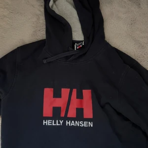 Svart hoodie från Helly Hansen - Säljer en svart hoodie från Helly Hansen med röd logga på bröstet. Den har en justerbar huva med grå insida och är perfekt för kyliga dagar. Passar både för vardag och avslappnade aktiviteter.
