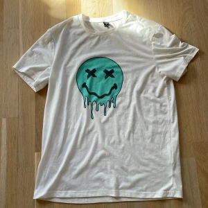 Vit t-shirt med tryck från Shein - Säljer en vit t-shirt från Shein med ett coolt tryck av ett smältande ansikte i turkos. T-shirten är kortärmad och gjord i en mjuk polyesterblandning. Perfekt för en avslappnad stil!