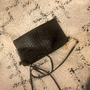 Svart axelväska med krokodilmönster - Snygg svart axelväska med krokodilmönster och dragkedja. Perfekt för att bära det viktigaste med stil. Den har en justerbar axelrem och en elegant design som passar till många tillfällen.