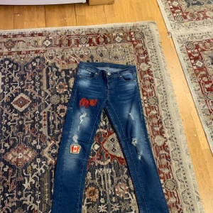 Blå jeans med broderier från Dsquared2 - Snygga blå jeans från Dsquared2 med coola broderier på framsidan, inklusive en röd text och en kanadensisk flagga. Jeansen har slitna detaljer och en klassisk femficksdesign. Perfekta för en trendig look!