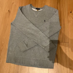 Grå sweatshirt från Ralph Lauren - Säljer en stilren grå sweatshirt från Ralph Lauren med rund hals och det klassiska logotypbroderiet på bröstet. Tröjan har långa ärmar och ribbade muddar vid ärmslut och nederkant. Perfekt för en avslappnad look.Säljer då den inte sitter så bra på mig använd fåtal gånger.