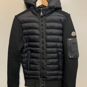 Svart jacka från Moncler - Snygg svart jacka från Moncler med quiltad framsida och huva. Jackan har långa ärmar och en dragkedja framtill. Det finns en broderad Moncler-logga på ärmen och en praktisk ficka med dragkedja.