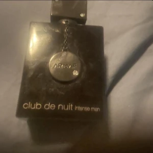 Club de Nuit Intense Man Parfym - Säljer en elegant och maskulin parfym från Armaf, Club de Nuit Intense Man. Flaskan är svart med en stilren design och har en metallkedja med en rund bricka. Perfekt för den som söker en intensiv och långvarig doft. Inspererad av creed aventus 