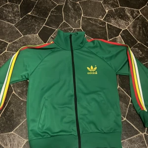 Grön träningsjacka från Adidas - Säljer en grön träningsjacka från Adidas med dragkedja och färgglada ränder på ärmarna. Jackan har Adidas-loggan i gult på bröstet och ryggen. Perfekt för en sportig look!