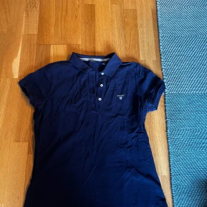 Mörkblå pikétröja från GANT - Säljer en klassisk mörkblå pikétröja från GANT med korta ärmar och knappar framtill. Perfekt för en stilren och avslappnad look.