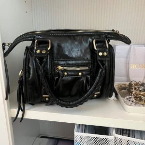 citybag, handväska - Snygg svart axelväska i skinn med flera detaljer. Väskan har en praktisk dragkedja och dekorativa nitar. Perfekt för att bära dina dagliga nödvändigheter med stil.
