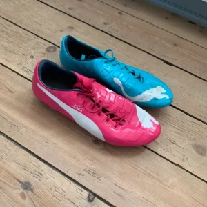 Puma EvoPower 1 Tricks - Säljer ett par unika fotbollsskor från Puma i olika färger. Den ena skon är rosa med vita detaljer och den andra är blå med vita detaljer. Skorna har snörning och är designade för att ge bra grepp på planen.