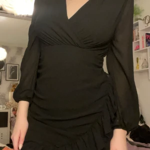 Svart omlottklänning med volang detalj  - Snygg svart omlottklänning med v-ringning och långa ärmar. Klänningen har volangdetaljer och en åtsittande passform som framhäver figuren. Perfekt för en kväll ute eller en speciell tillställning. Lapp bortklipp men vet är köpt från chiquelle  S 