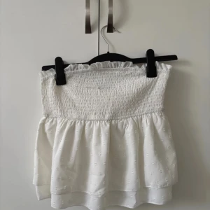 Vit axelbandslös topp från Shein - Säljer en söt vit axelbandslös topp från Shein. Toppen har en smockad överdel och ett lätt volangfall nedtill. Perfekt för varma sommardagar! Mindre i storleken.