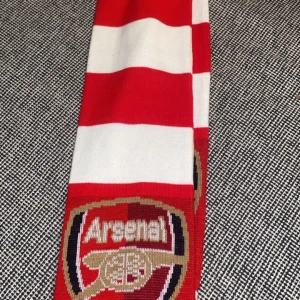 Röd och vit randig halsduk från Arsenal - Snygg halsduk i rött och vitt med Arsenal-logga. Perfekt för att visa ditt stöd på matchen! Halsduken har fransar i ändarna och är tillverkad i ett mjukt material.