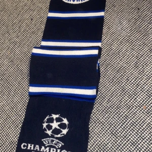 Blå och vit halsduk med Champions League-motiv København - Snygg halsduk i blått och vitt med Champions League-motiv och fransar i ändarna. Perfekt för fotbollsfans som vill visa sitt stöd. Den har ett randigt mönster och är märkt med UEFA Champions League.