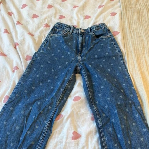 Blå jeans med hjärtmönster från H&M - Snygga blå jeans från H&M med ett gulligt hjärtmönster. De har en hög midja och en justerbar passform för extra komfort. Byxorna har en bred stil och är perfekta för en avslappnad look. Klassisk knapp och dragkedja framtill. Storlek 146