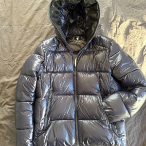 Mörkblå pufferjacka från Save The Duck - Snygg svart pufferjacka från Save The Duck med glansig finish och huva. Jackan är 100% animal free och fylld med Plumtech syntetdun. Perfekt för kalla dagar med dragkedja framtill och praktiska fickor. Nypris 4000kr