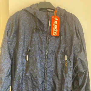 Blå vindjacka från Superdry - Snygg blå vindjacka från Superdry med dragkedja och justerbar huva. Jackan har ett diskret mönster och praktiska fickor med dragkedjor. Perfekt för blåsiga dagar.