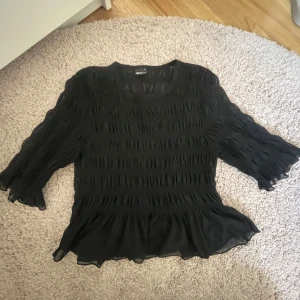 Svart blus med volangdetaljer - Säljer en elegant svart blus från Gina med volangdetaljer. Blusen är perfekt för en stilren look. Materialet är lätt och luftigt, vilket gör den bekväm att bära.