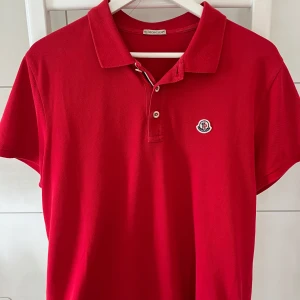 Röd pikétröja från Moncler - Säljer en fett unik röd pikétröja från moncler, den har fungerande qr kod och är köpt på garmsmarket så de finns bevis på att den är äkta. Storlek är XL men passar L. Den har dock ett par mindre hål på ryggen som knappt syns men också går enkelt o laga!! Bara att höra av er vid frågor🙌