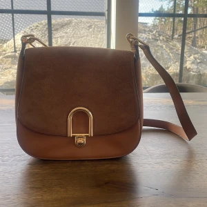 Brun axelväska från Michael kors  - Snygg brun axelväska från Michael Kors i en kombination av skinn och mocka. Väskan har en elegant guldfärgad låsdetalj framtill och justerbar axelrem. Perfekt för att ge din outfit en stilfull touch.