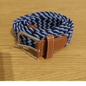 Blått flätat bälte med läderdetaljer - Snyggt flätat bälte i blått och ljusblått med bruna läderdetaljer och ett silverfärgat spänne. Perfekt för att ge en stilren touch till din outfit. Passar bra till både jeans och chinos.