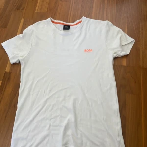 Vit t-shirt från Boss - Säljer en stilren vit t-shirt från Boss med en liten logga i orange på bröstet. T-shirten har en klassisk rund halsringning och korta ärmar. L men passar M